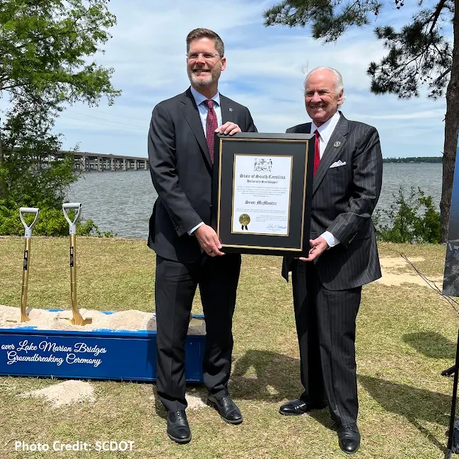 Two men in suit holding a framed proclamation next to a ceremonial sand pit with two shovels in front of water and trees. Photo Credit: SCDOT