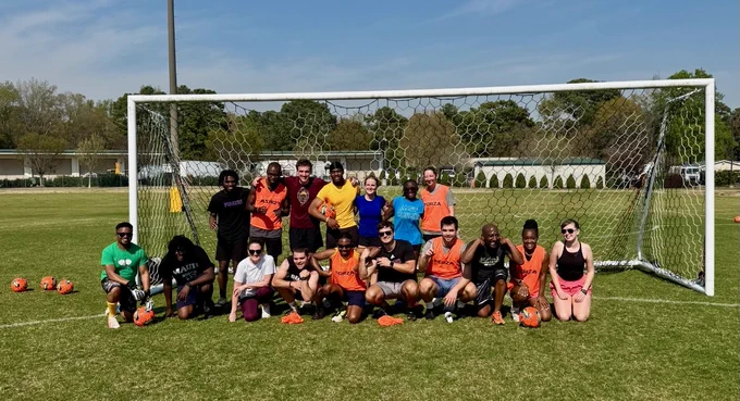 A group of people are posing in front of a soccer goal on a grassy field. They are dressed in athletic attire, some wearing orange pinnies. Several soccer balls are scattered on the ground in front of them. 