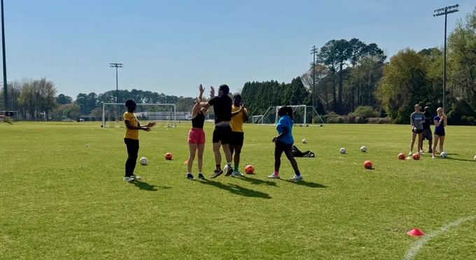 A group of people on a grassy soccer field, some wearing orange vests. They are surrounded by soccer balls, with goalposts in the background and trees lining the field.
