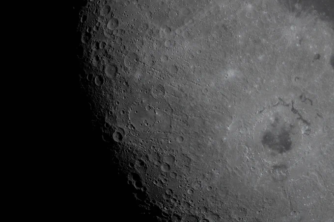 A close-up view of the Moon, filling most of the frame. Along the left side of the image, the terminator divides the illuminated and darkened sides of the Moon. Just before the terminator, the lunar surface transitions from the smoother near side to the bumpier, more cratered far side. From this perspective, the distinction between the two is clearer. The craters on the far side create a bumpy, textured surface. Near the lower right hand corner of the image, a large circular basin has darker mottling near its center. Credit: NASA