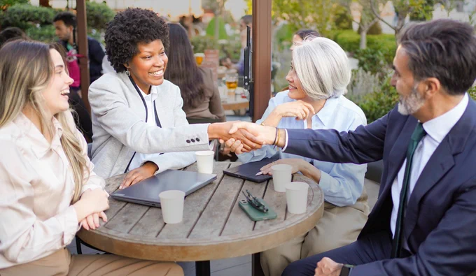 A man in business clothing shaking hands with a woman in business clothing with 2 other women in business clothes sitting at the same table.