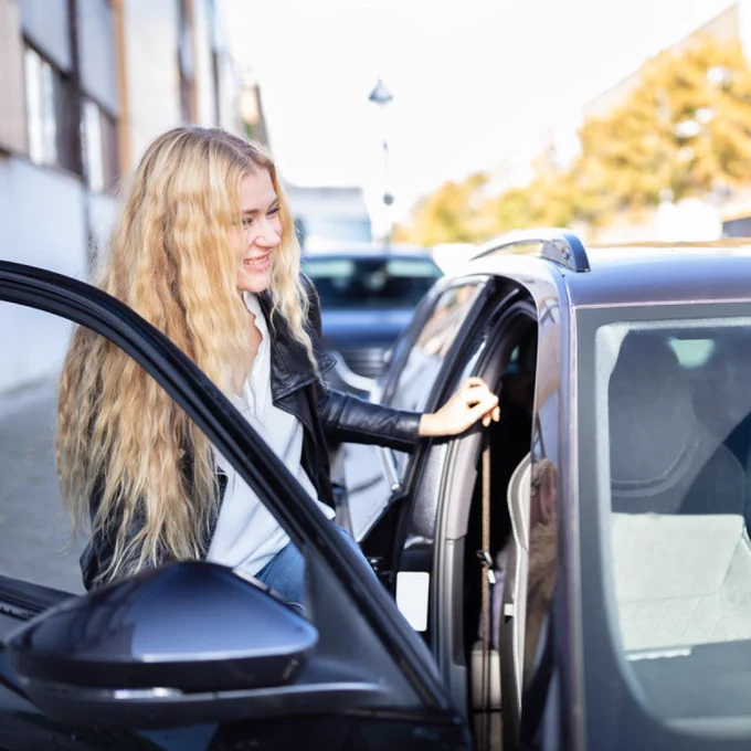 Woman with long blonde hair opening the rear door of a black car parked on a city street during daytime.