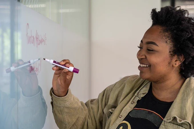Smiling Black participant in a green jacket and black t-shirt, whiteboarding something unreadable
