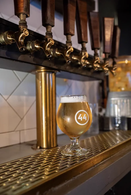 A glass of beer sits on a metal bar counter beneath a row of wooden taps. The glass has a "44" logo on it.