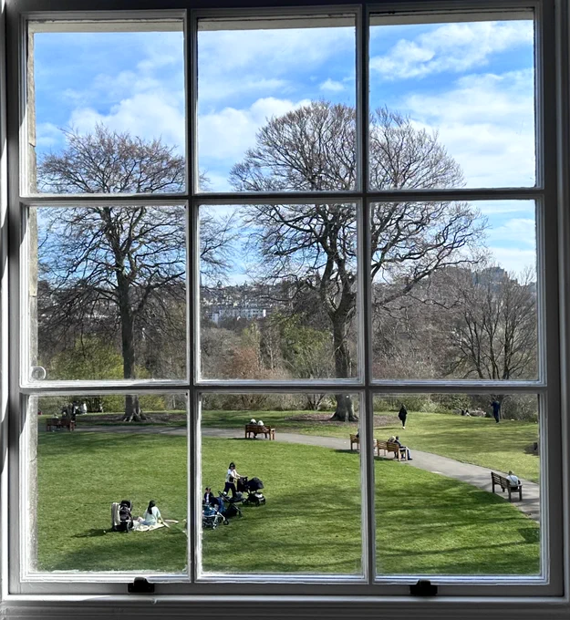 View through a sash window towards people sitting on the grass, leafless trees, Edinburgh in the background and a blue sky with white clouds.