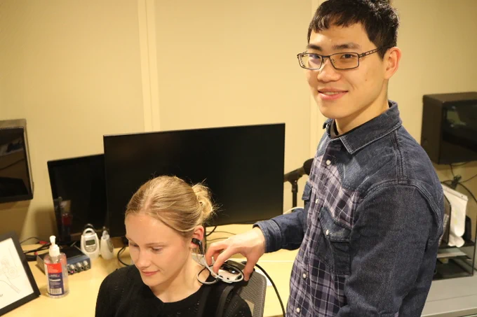 Two students in the Auditory and Mechanics Perception Lab performing a routine hearing exam. They are in a dimly lit cream-colored sound booth. The one on the right is facing the camera and smiling, while the one receiving the exam is facing left with a soft smile.