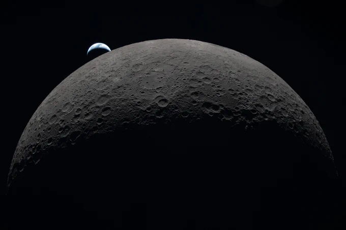 Alt: Earth setting behind the Moon. The gray half circle of the Moon, pocked with craters, fills the majority of the image, brightening from darkness at the bottom to light gray at the top, stark against the blackness of space. A much smaller blue and white crescent of Earth, oriented like a frown, appears to touch the edge of the Moon at one tip near the 11 o\u2019clock region of the image.