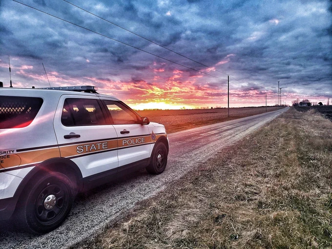 A State Police SUV is parked on the side of a rural road. The sun is setting in the distance, casting vibrant orange and pink hues across the cloudy sky. The fields on either side of the road are expansive and devoid of structures, with utility poles lining the road.