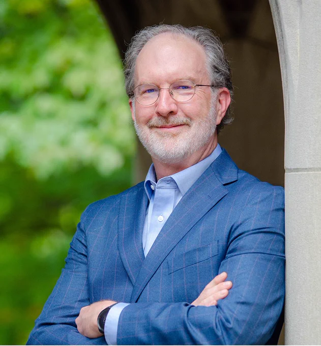 A man with glasses leaning against a concrete archway. He is wearing a blue button-up shirt with a blue blazer jacket.