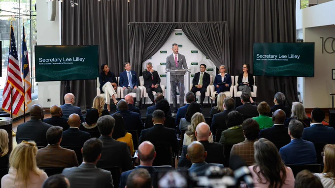 A group of people seated in rows facing an indoor stage with a podium. The person at the podium, Secretary Lee Lilley, is speaking.