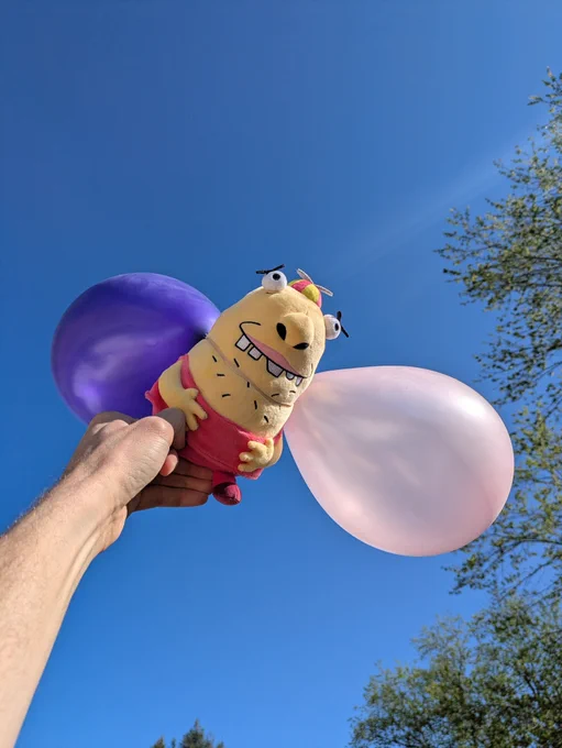 The Beans plushie is held up to the sky with a couple of balloons attached.