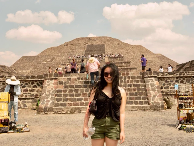 A person stands smiling in front of the Pyramid of the Sun at Teotihuacan, holding a water bottle. Other visitors can be seen on the pyramid and nearby. Souvenir stalls are visible on the sides.