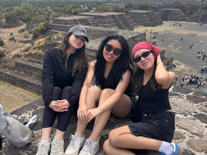 Three people sitting and smiling in front of the ancient pyramids of Teotihuacan. They are on a stone platform with the vast archaeological site and tourists visible in the background.