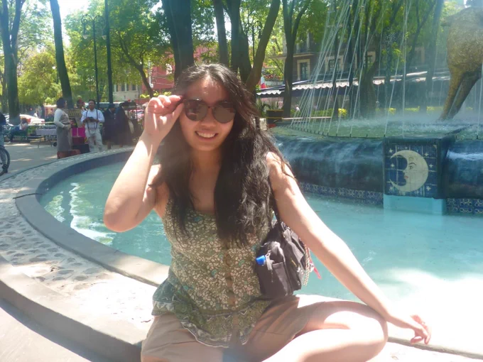 A fountain in a sunny outdoor setting with trees and pedestrians in the background.