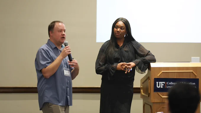 Two people presenting at a podium in front of a crowd with a presentation behind them.