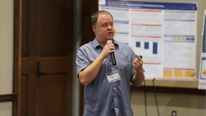 A person holding a microphone and speaking to an audience in front of a poster board.