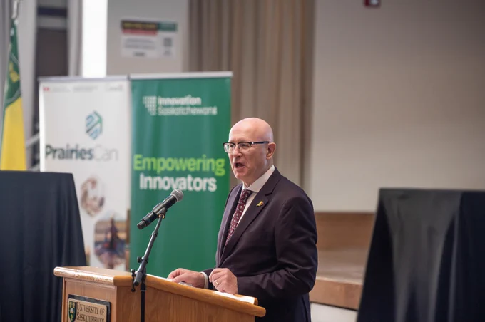 Speaker addressing an audience from a podium at a University of Saskatchewan venue, with Innovation Saskatchewan and PrairiesCan banners behind him, highlighting collaboration and investment in innovation, research and economic development in Saskatchewan.