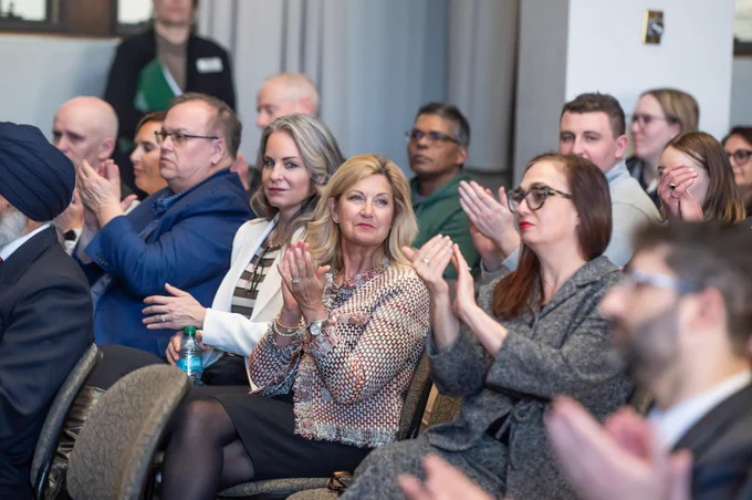 Audience members seated at an innovation and research event in Saskatchewan applaud during a presentation, with professionals from government, academia and industry shown listening and engaging in a formal indoor venue.