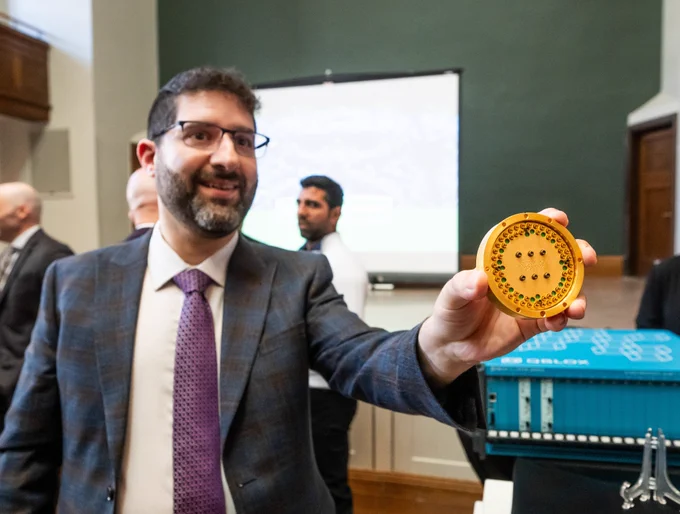 Man in a suit holds up a circular quantum computing component during a technology demonstration at an event, with research equipment and attendees visible in the background, highlighting advances in quantum science and innovation in Saskatchewan.