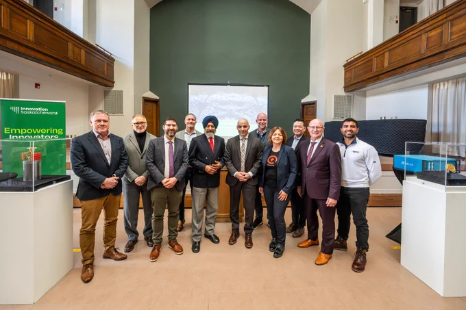 Group of government, academic and industry leaders standing together in a large hall during an event, with display cases and a presentation screen in the background, highlighting collaboration and innovation in Saskatchewan’s research and technology ecosystem.