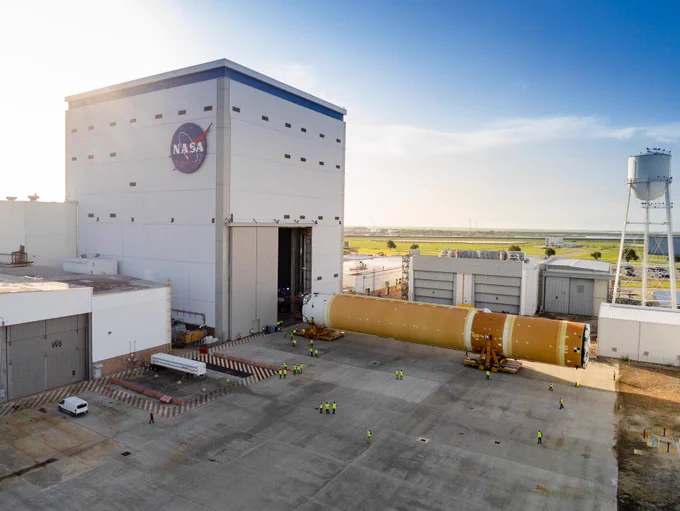 NASA and Boeing prepare the butterscotch-orange-colored SLS (Space Launch System) rocket core stage for shipment at NASA\u2019s Michoud Assembly Facility in New Orleans. The core stage is on its side on two orange structures that hold it above the ground. The core stage is just in front of a tall rectangular building with a NASA meatball logo on the side facing us. The building's massive doors are open. Credit: NASA/Eric Bordelon