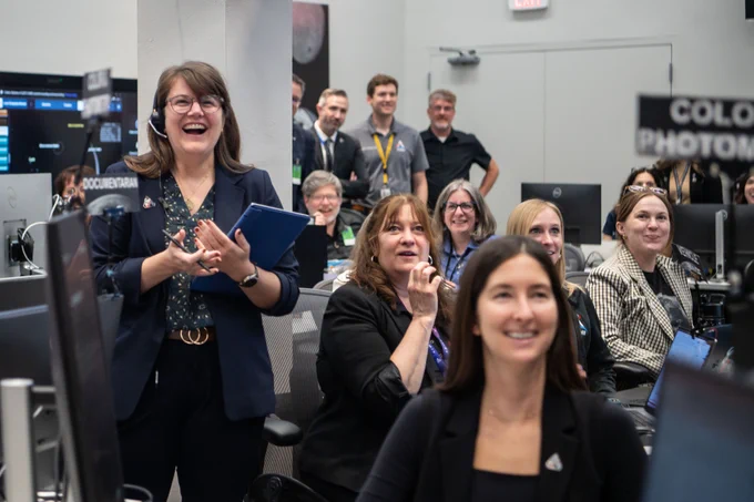 A picture of a group of women with joyous reactions on their faces as they look up at something off screen. They wear business attire and have big smiles on their faces. A small group of men is in the background, out of focus. 