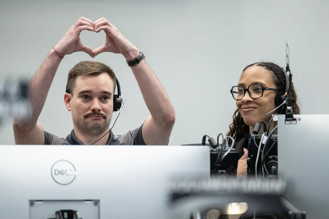 A picture of a man and woman standing behind large computer monitors. The man, on the left, is lifting his hands in the shape of a heart above his head. The woman, on the right, has a large smile on her face. 