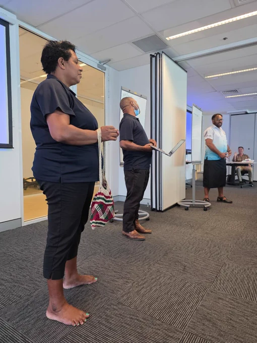Participants presenting at the front of the room, one holding a laptop and another carrying a woven bag, engaged in a discussion.