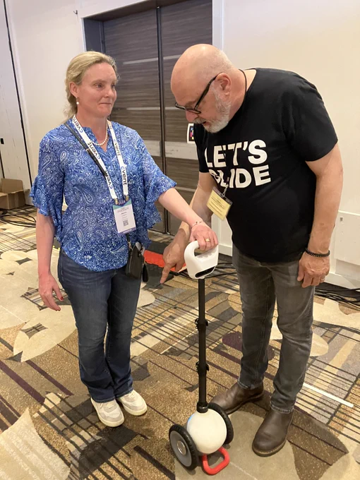 Jessie stands with Glide, a compact two-wheeled mobility device with a rounded white base and a vertical handlebar, holding it as it rests on the floor ready to guide her movement.