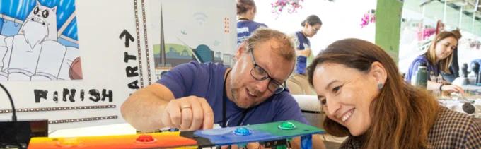 Two people at a workshop table assembling an electronic project with colourful components and wires. One person is adjusting the device while the other smiles and watches closely.