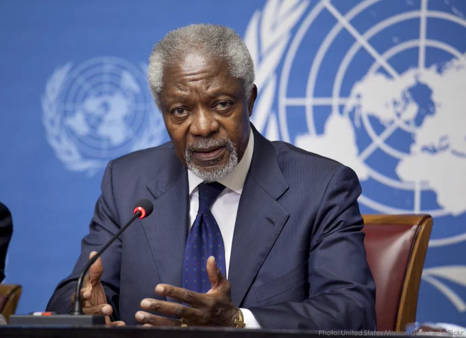 Kofi Annan sits at a table, speaking, with a UN logo background.