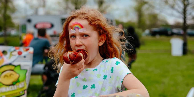 Young girl with curly red hair enjoying a hotdog at an outdoor picnic table with painted rocks in front of her.