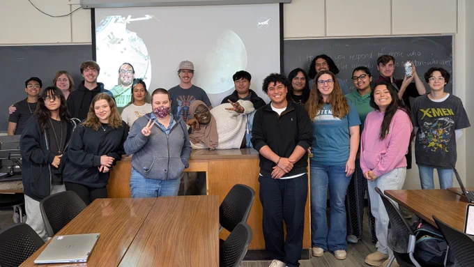 A picture of Dr. Purvis' Space Physics class posing in front of a projector screen that is showing the Artemis II mission livestream.