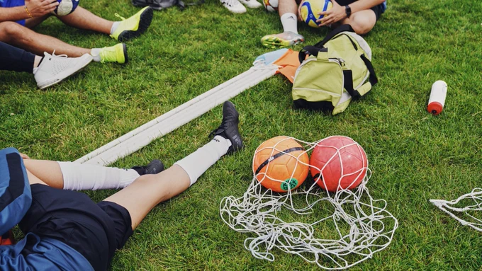People sitting on grass with sports gear, including footballs, a net, and a backpack.