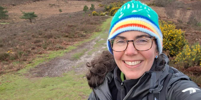 A New Forest National Park Authority ranger in outdoor gear and a colorful beanie is smiling, holding a camera in a natural landscape with grassy and bushy terrain. 