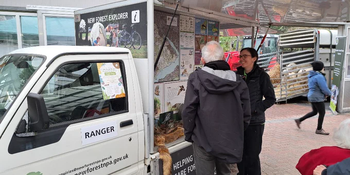 A New Forest National Park Authority ranger is standing next to a New Forest NPA information van, speaking with a visitor at the New Forest Marque Spring Market roadshow event at Everton Nurseries. The van displays a "New Forest Explorer" sign and informational posters. The scene is in an outdoor area with a few other people walking in the background.