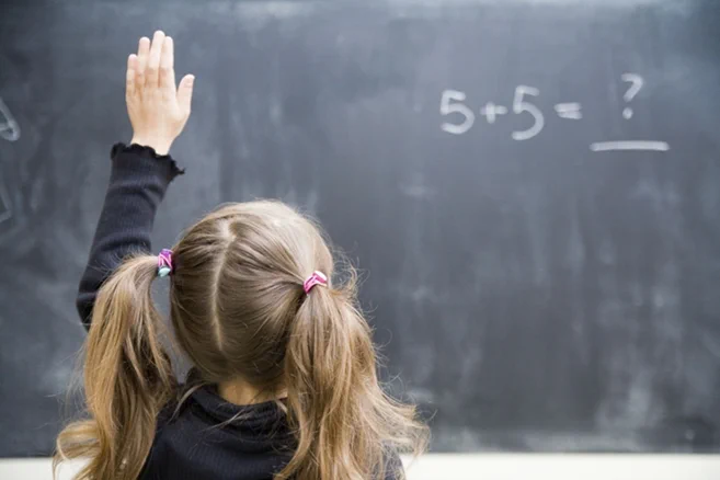 Photograph of a young girl with pigtails raising her hand in front of a chalkboard displaying the math problem "5   5 = ?". 