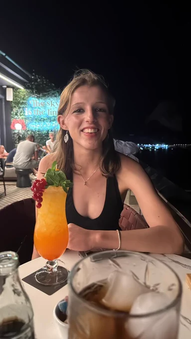 Rose is a white woman in her twenties with shoulder-length blonde hair. She sits in a restaurant outdoors with an orange cocktail.