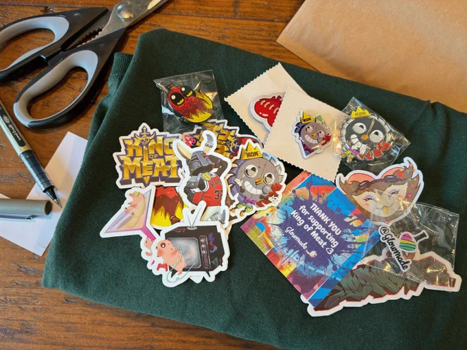 A collection of colourful King of Meat stickers and enamel pins spread across a dark green t-shirt, with scissors, a pen and brown envelopes in the corners of the frame.