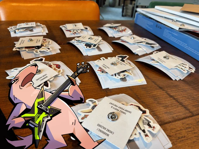 Multiple stacks of enamel pins featuring various designs neatly arranged on a wooden table, with a cartoon Artvark playing an electric guitar-axe in the foreground.
