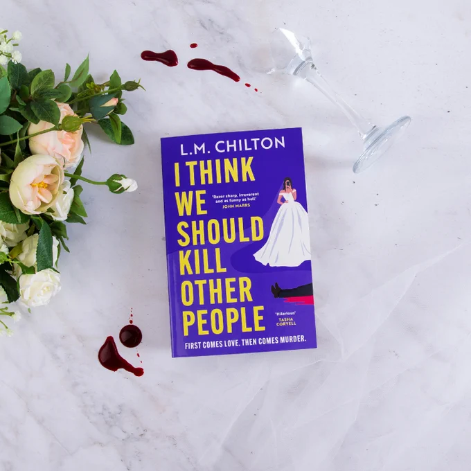 A book titled "I Think We Should Kill Other People" by L.M. Chilton lies on a marble surface. Nearby are a spilled glass with red liquid and a bouquet of white and peach roses.