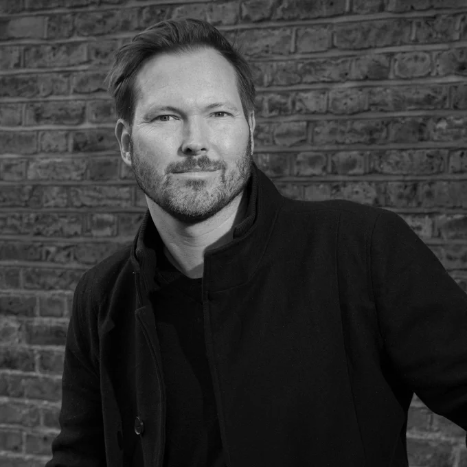 A black and white photograph of the author L.M. Chilton against a brick wall. 
