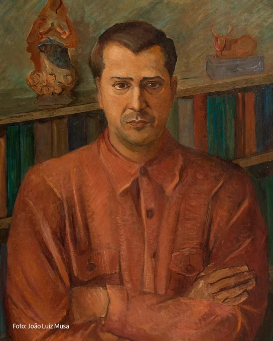 Fotografia da obra “Retrato de Luís Martins I”, de Tarsila do Amaral, que destaca um homem branco de cabelos castanhos. Ele usa uma camisa alaranjada e está de braços cruzados. Ao fundo, há uma estante com livros.