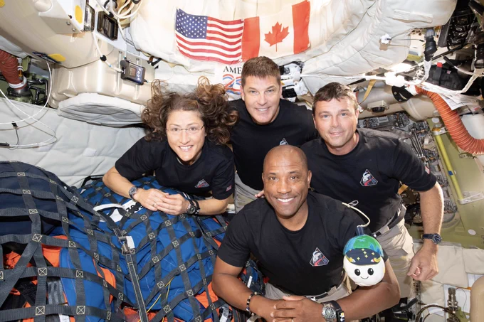 Four astronauts float close together inside a spacecraft. They are wearing matching black shirts, and the area around them is filled with equipment, cables, and storage bags. A large bundle wrapped in blue straps sits in the foreground. A small, round plush toy is attached to the arm of one crew member. Behind them, American and Canadian flags are attached to the wall along with other gear.