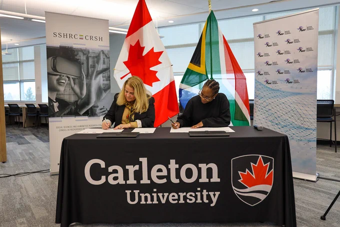 Two women in business attire signing documents in front of the Canadian and South African flags. Tablecloth is branded with the Carleton logo.