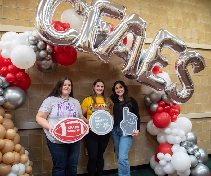 3 current students standing in front of photo backdrop posing for a picture holding CFAES props. 
