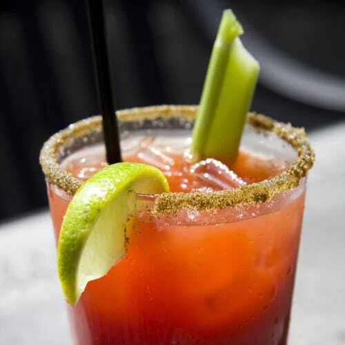 A close-up of a Caesar cocktail in a glass with a salted or spiced rim. The drink is reddish-orange and filled with ice. A lime wedge is hooked onto the rim, and a celery stalk stands upright inside the glass. A black straw is also visible. The background is softly blurred, keeping focus on the drink.
