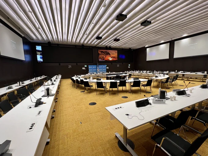 A large conference room with numerous white tables arranged in a U-shape. Each table has microphones and nameplates. A large screen displays a colorful abstract art piece at the front. The room is well-lit, with a patterned ceiling and two large projection screens on the side walls.