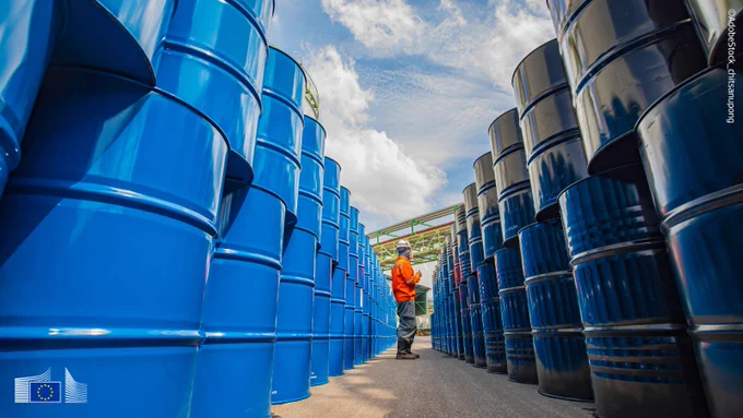 Blue oil barrels on both sides of a narrow path, where a person in orange jacket and white helmet verifies something. 