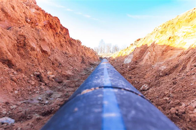 Large black pipeline laid in a deep trench surrounded by reddish soil under a clear blue sky.